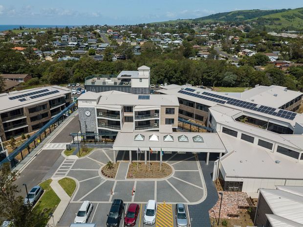 Aerial view of a contemporary multi-building institutional or commercial complex with light-coloured metal roofs, extensive rooftop solar panels, glazed façades, a covered central entrance, landscaped courtyards, parking areas, and surrounding suburban housing and rolling green hills.