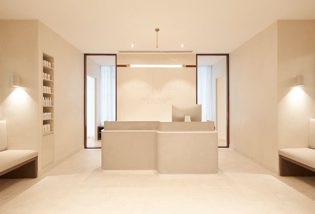 Minimalist, symmetrical reception or concierge area with a central pale stone or plaster desk, built-in upholstered benches along both walls, recessed shelving, warm cream finishes, and glass-framed openings at the rear. Soft concealed and wall lighting creates a calm, refined atmosphere.
