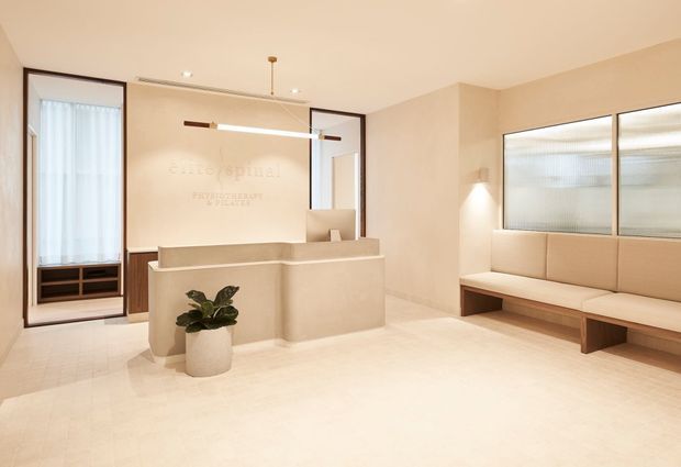 A bright, minimalist reception area with a warm neutral palette. A sculptural cream reception desk sits centrally beneath a slim linear pendant light, with built-in upholstered bench seating along the right wall. Floor-to-ceiling glazing and dark-framed doors add contrast, while a small potted plant provides a natural accent.