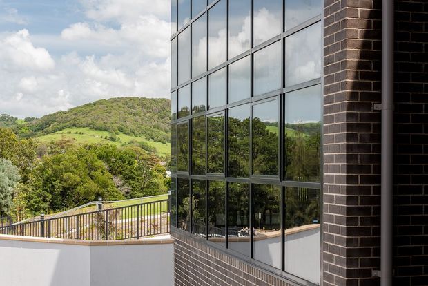 Exterior architectural view of a contemporary building featuring a tall grid of reflective floor-to-ceiling glazing set beside dark brick masonry. A balcony or terrace with metal railing is visible in the foreground, while green rolling hills are reflected in and seen beyond the facade.