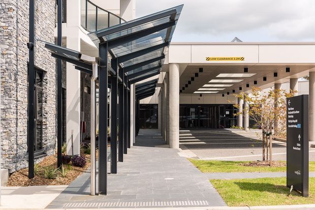 A contemporary commercial building entrance with a covered pedestrian walkway, angular glazed canopy, dark steel supports, pale rendered walls, and a textured stone-clad facade. The entrance canopy leads toward a covered drop-off area with columns, directional signage, landscaped lawns, and young trees.