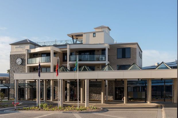 Exterior view of a multi-storey contemporary civic or commercial building with a broad covered entry canopy, slender columns, glass-fronted balconies, brick and stone-clad walls, rooftop structures, and several flagpoles. A clock is mounted on the left facade, and the building sits within a paved landscaped forecourt.