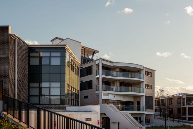 Exterior architectural photograph of a contemporary multi-storey residential or mixed-use building complex. The main building combines dark brick masonry, extensive dark-framed glazing, curved projecting balconies, white rendered walls, and a flat/low-pitched roof profile. A sloping access path and black metal fencing occupy the foreground, with adjoining modern buildings visible to the right.