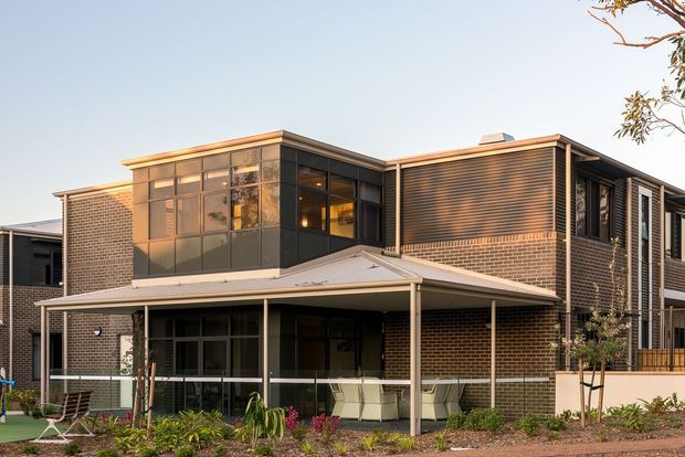 Exterior view of a contemporary multi-storey brick residence with a broad covered verandah, extensive dark-framed glazing, projecting upper-level volumes, and horizontal dark cladding. Landscaping and outdoor furniture sit in the foreground.