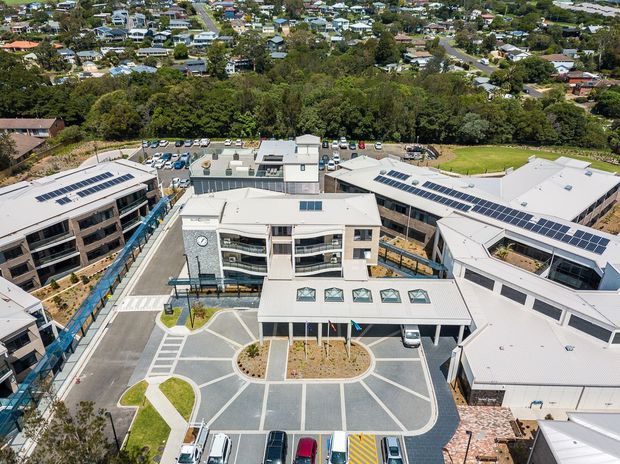 Aerial view of a contemporary multi-building development with light-coloured metal roofs, extensive rooftop solar panels, landscaped pedestrian areas, internal driveways, and car parking. A dense suburban neighbourhood and mature vegetation surround the complex.