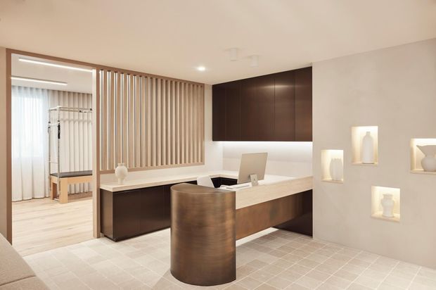 A refined contemporary reception or entry area featuring a dark timber built-in desk and cabinetry, a curved timber-fronted counter, pale tiled flooring, and a vertical timber-slat divider screening a bright adjoining room. Recessed wall niches display decorative objects, while soft integrated lighting creates a warm, polished atmosphere.
