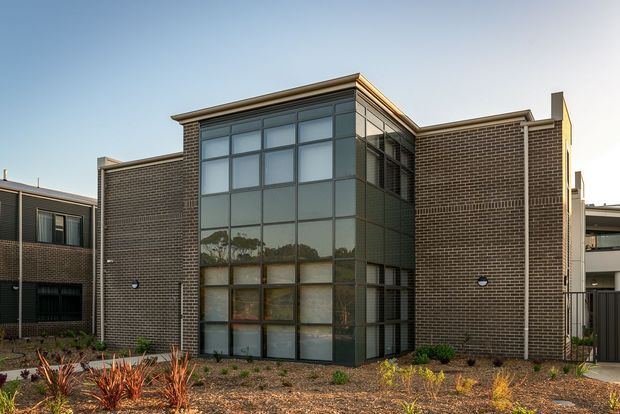 Exterior view of a contemporary multi-storey brick building with a prominent floor-to-ceiling glazed corner volume, flat rooflines, and a low-maintenance landscaped frontage. The facade combines dark brown face brick with dark metal-framed glazing and restrained modern detailing.