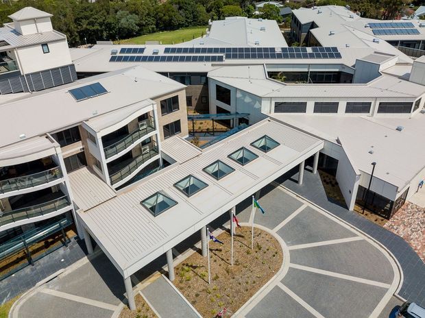 Aerial view of a large contemporary institutional or commercial complex arranged around landscaped courtyards and paved pedestrian areas. The buildings feature pale metal pitched and low-slope roofs, extensive skylights, solar panels, dark framed glazing, covered walkways, balconies, and a central flagpole forecourt.