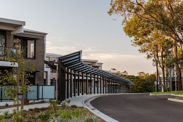 Contemporary multi-unit residential development viewed from a landscaped driveway, featuring dark brick facades, flat rooflines, extensive glazing, and a long sculptural curved covered walkway or carport structure with dark steel framing and translucent roofing. Mature trees and planted garden beds surround the paved access road.