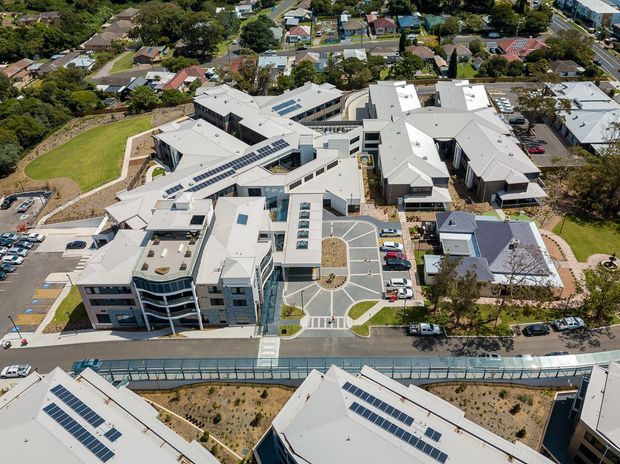 Aerial view of a large contemporary institutional or commercial campus with multiple interconnected low-rise buildings, pale pitched and flat roofs, extensive rooftop solar panels, landscaped courtyards, internal roads, parking areas and surrounding suburban housing.