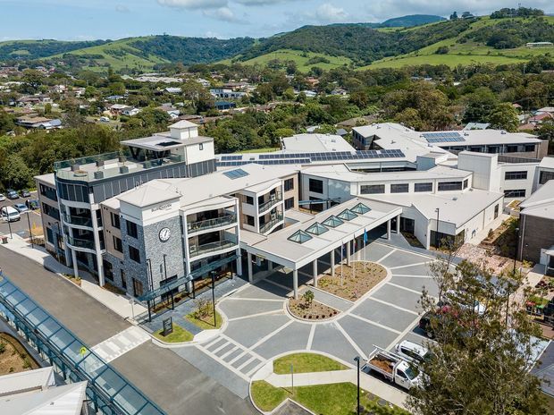 Aerial view of a contemporary multi-building institutional or commercial complex arranged around landscaped paved courtyards. The buildings have predominantly white and grey facades, extensive flat roofs with solar panels, rooftop equipment, glazed links, and a clock tower feature. Hilly green suburban terrain is visible in the background.