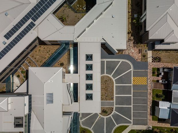 Top-down aerial view of a contemporary multi-building development with light metal roofs, extensive rooftop solar panels, landscaped courtyards, internal pedestrian paths, and a patterned paved driveway or parking area. The complex features angular roof forms, skylights, and integrated outdoor circulation spaces.