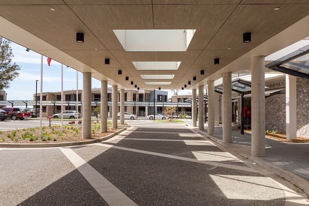 Wide-angle exterior view of a covered drop-off or pedestrian forecourt in a contemporary institutional or commercial complex. A large timber-toned soffit with rectangular skylights and recessed cylindrical lights is supported by regularly spaced round concrete columns. Paved circulation areas, landscaped beds, stone-clad walls, and glazed building wings are visible beyond.