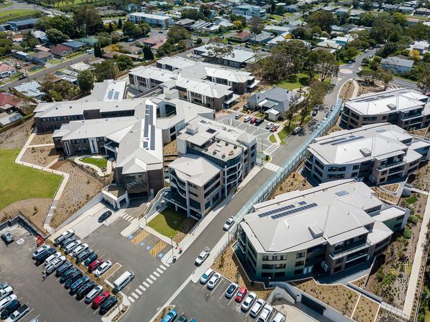 Aerial view of a contemporary multi-building development, likely a university or institutional campus, with several light-coloured pitched and flat-roofed buildings, landscaped courtyards, internal roads, pedestrian paths, and extensive parking. Solar panels are visible on some roofs, and the surrounding suburban neighbourhood is visible beyond the site.