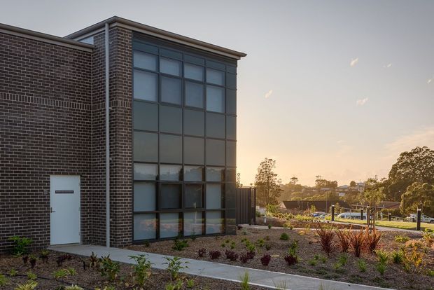 Modern two-storey brick building exterior with a tall glazed curtain-wall section, flat rooflines, and a landscaped garden frontage. The image is photographed at sunset, with a pathway and low planting leading toward the building entrance.