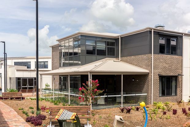 Exterior view of a contemporary multi-storey brick and glass building with a projecting low-pitched roof, extensive glazing, dark horizontal cladding and an elevated covered terrace or entry area. The landscaped foreground includes paved paths, garden beds and outdoor seating. The building appears institutional or commercial rather than residential.