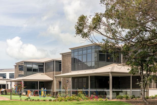 Exterior view of a contemporary multi-wing brick building with broad covered verandas, extensive dark-framed glazing, louvred screens, and low-pitched metal roof forms. A landscaped garden and playground occupy the foreground, with mature trees partially framing the building.