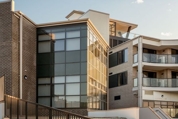 Exterior view of a contemporary multi-storey residential apartment building, with dark brick facades, extensive multi-level glazing, projecting balconies, metal balustrades, and a mix of flat and low-pitched roof forms. The image is focused on the building facade and shared exterior access areas.