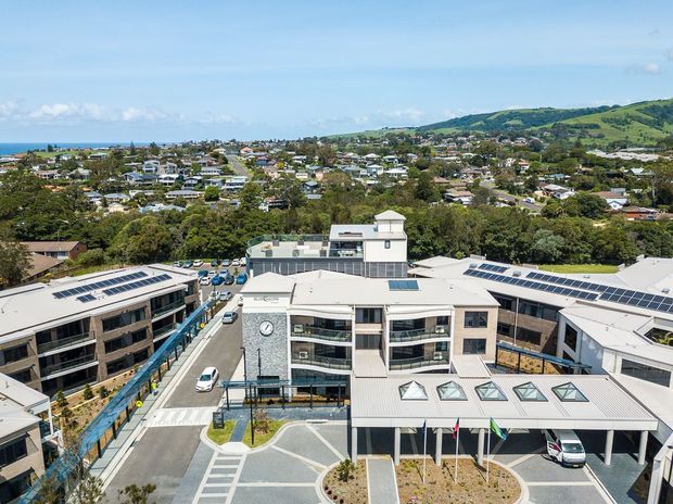 Aerial view of a contemporary multi-storey commercial or institutional complex with pale metal roofs, extensive rooftop solar panels, glazed balconies, landscaped courtyards, and a formal entrance drive. A dense suburban neighbourhood, green hills, and the ocean are visible beyond the development.