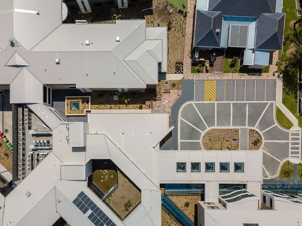 Aerial top-down view of a contemporary multi-building complex with several light metal-roofed structures, rooftop courtyards and skylights, solar panels, landscaped internal areas, and a geometric paved parking and circulation zone. The image focuses on the overall site layout and building roofs rather than a single interior space.
