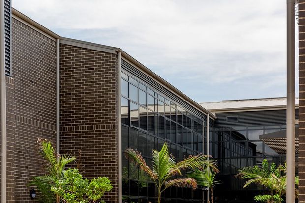 Exterior architectural view of a contemporary multi-wing building centred on a glazed connecting corridor or atrium. Dark brown brick walls, charcoal metal framing and extensive floor-to-ceiling glazing define the facade, with palms and planting in the foreground.