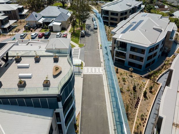Aerial view of a contemporary multi-building urban development, showing flat and low-pitched rooflines, rooftop solar panels, landscaped courtyards, internal roads, pedestrian crossings, and a prominent covered pedestrian walkway or glazed canopy running through the site.