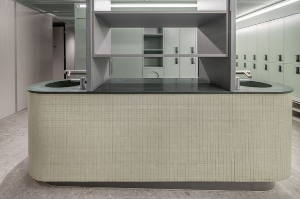 A contemporary commercial washroom interior featuring a large curved vanity island clad in small pale mosaic tiles, with dark stone or composite benchtops, integrated basins, and chrome tapware. Mirrored panels and geometric open shelving sit above the vanity, while pale built-in cabinetry and multiple wall-mounted fixtures extend into the background. The space has a cool, refined, highly symmetrical appearance.