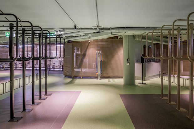 A large contemporary commercial interior resembling a transit or public facility, with exposed services and ductwork, pale green structural columns, tiled service enclosures, and rows of dark metal tubular railings or barriers. The floor uses broad geometric sections of muted pink, pale green, and dark brown, creating a graphic, functional atmosphere.