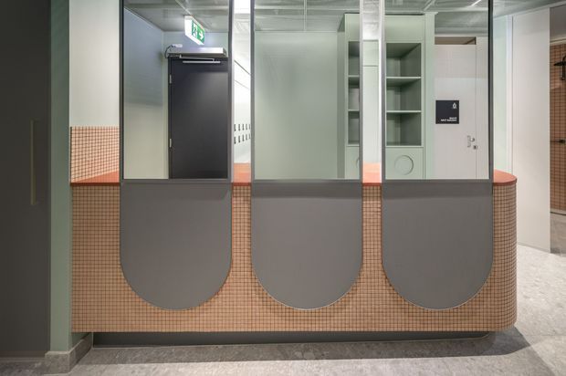Contemporary commercial interior featuring a curved reception or service counter clad in small peach-toned mosaic tiles, with three rounded grey privacy panels and a grey stone-like floor. Behind the counter are glazed partitions, open shelving, a dark door, and a visible exit sign. The space has a clean, refined institutional or hospitality fit-out with muted green and grey tones.