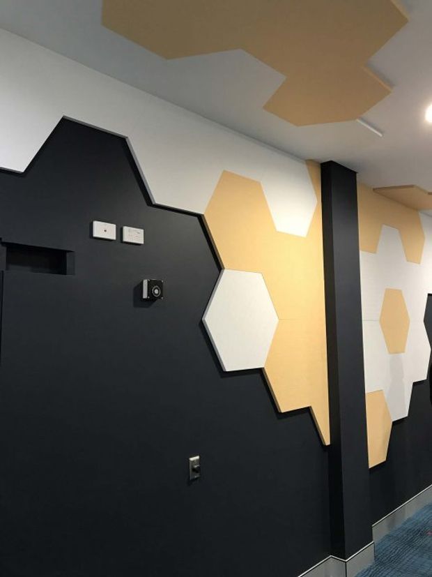 Interior view of a bold, geometric feature-wall installation. Large interlocking black, white, and warm ochre polygonal panels extend across the walls and ceiling, with a black structural column dividing the space. Electrical outlets and a small wall-mounted device are visible, along with a dark patterned carpet floor and recessed ceiling lighting. The space has a graphic, contemporary commercial or media-oriented character.