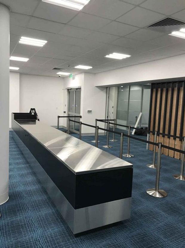 A clean, modern commercial interior resembling an airport or transport terminal check-in and queuing area. The space features a long black reception or service counter with a brushed stainless-steel top and base, blue patterned carpet, white walls, suspended grid ceiling with recessed fluorescent lighting, glass partitions, and black queue barriers with stainless-steel posts. A vertical timber slat screen forms a feature partition at the rear.