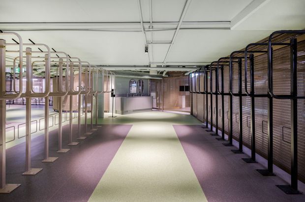 A large contemporary commercial interior with rows of enclosed workstation or display structures lining both sides of a broad central aisle. The space features exposed ceiling services, polished concrete or smooth flooring in muted purple and pale green sections, translucent and timber-toned partitions, and a reception or service counter at the far end. The overall atmosphere is modern, spacious, and softly illuminated.