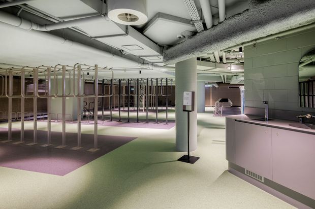A large open-plan commercial interior with exposed services and ducts across the ceiling, pastel green and mauve flooring, structural columns, rows of rounded metal-framed partitions or display fixtures, and a curved reception or service counter in the foreground. The space has a contemporary industrial fit-out with soft, colourful finishes and bright diffuse lighting.
