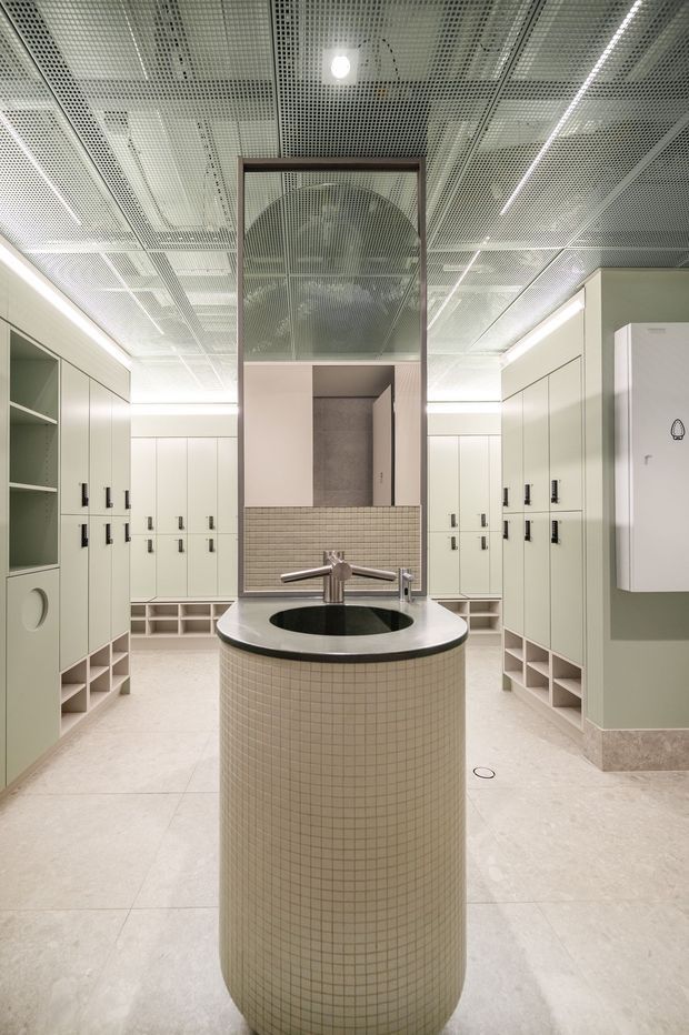 A contemporary commercial locker-room or changing-room interior with pale sage-green built-in lockers, open cubby storage, and a central cylindrical tiled hand-washing basin with a dark stone rim and sculptural faucet. The space has a light stone or terrazzo floor, a suspended perforated acoustic ceiling with linear lighting, and a large central mirror or glazed screen framed in dark metal.