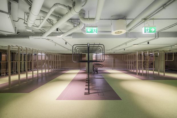A large, open interior with extensive exposed mechanical services and white-painted pipework across the ceiling. Repetitive metal tubular frames and rail-like structures form organised rows throughout the space, with muted pink, green, and grey floor zones and illuminated emergency exit signs. The utilitarian infrastructure and open plan suggest a commercial or institutional facility.