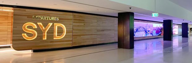 Wide interior view of a contemporary airport departures area, featuring a large curved timber-clad wall with illuminated “SYD” signage, polished light flooring, dark structural columns, and purple-blue illuminated digital artwork or wayfinding displays.