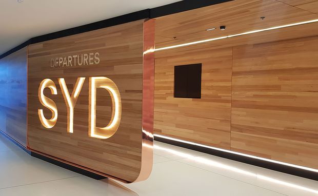 Interior airport wayfinding and departures signage featuring large illuminated “SYD” lettering and “DEPARTURES” text, set within a warm timber-clad wall. Linear integrated lighting, black trim, and a clean contemporary commercial fit-out define the space.