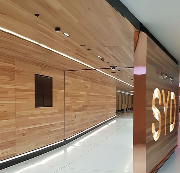 A polished commercial interior corridor or lobby with extensive light timber wall and ceiling panelling, glossy white tiled flooring, integrated linear LED lighting, dark trim details, and a large illuminated sculptural sign at the right. A glazed partition reflects the timber-lined passage and recessed lighting, creating a refined contemporary workplace or hospitality entrance.