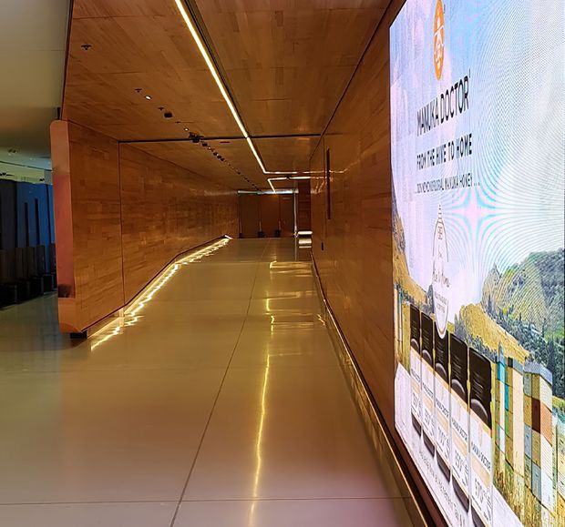 A polished commercial interior corridor or exhibition passage with warm timber wall and ceiling panelling, a glossy light-coloured tiled floor, integrated linear LED lighting, and a large illuminated digital display wall presenting project or product information. The space appears modern, refined, and gallery-like.