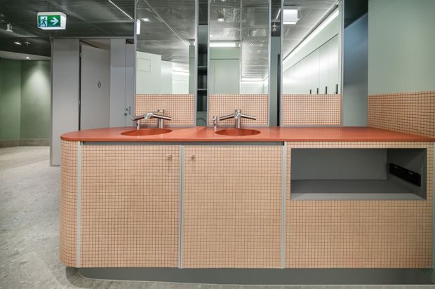 A contemporary commercial washroom interior with a long tiled vanity featuring two integrated basins, wall-mounted faucets, open storage, and matching pink-beige mosaic tile splashbacks. Large mirrors, pale green-grey wall panels, exposed ceiling services, and an accessible exit sign are visible.