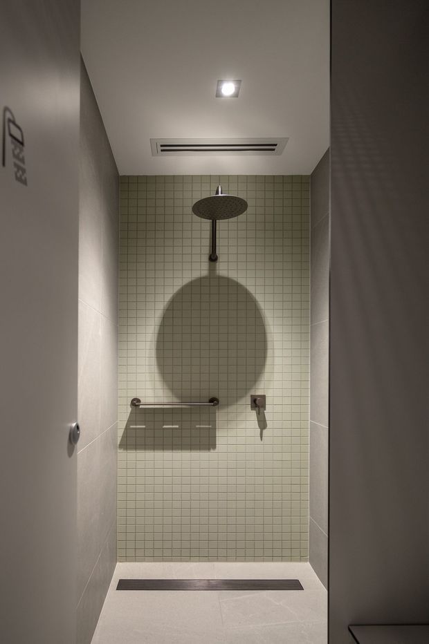 Compact contemporary walk-in shower with pale square mosaic tiles on the feature wall, larger light stone-look wall and floor tiles, a dark ceiling-mounted rain shower, recessed ceiling light and linear floor drain. Minimal dark metal fixtures and a shower bench or shelf create a clean, understated fit-out.