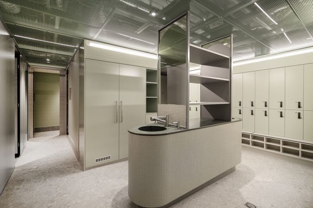 A contemporary commercial locker-room or changing facility with a central curved vanity island, integrated round basin, shelving, large white storage cabinets and extensive lockers. The space features a perforated metal ceiling, pale tiled or terrazzo-like flooring, white built-in joinery, and a muted grey-beige palette with reflective surfaces and bright linear lighting.