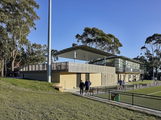 Exterior view of a contemporary multi-level community or institutional building set within a landscaped, gently sloping site. The building features a large flat projecting roof canopy, extensive glazed façades, metal screening and balustrades, pale masonry walls, and an adjacent fenced sports court or recreation area. Mature eucalyptus trees surround the site.