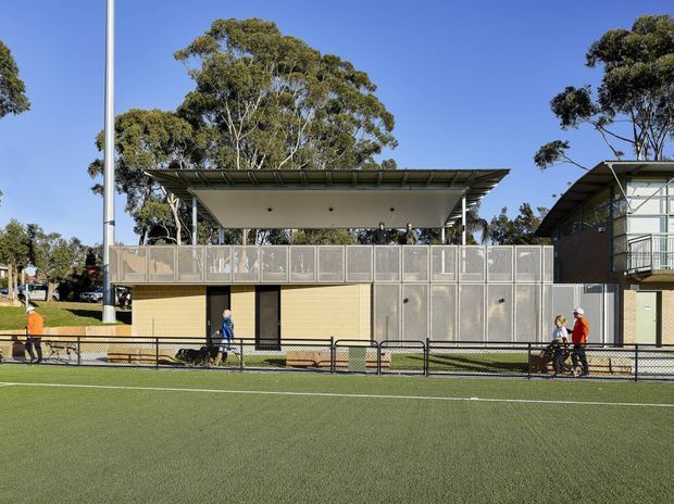 Exterior view of a contemporary low-rise sports or recreational facility beside an artificial turf field. The building has a flat, broad overhanging roof, exposed steel supports, perforated metal screening and railings, pale yellow brick or panel walls, and large glazed areas. A neighboring modern building and mature trees are visible in the background.