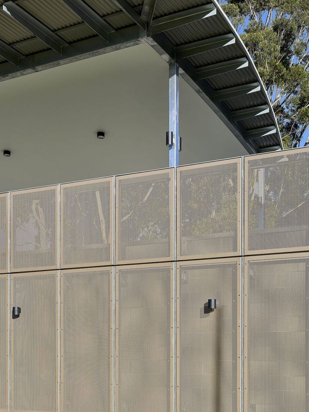 Close-up exterior view of a contemporary building facade beneath a projecting corrugated metal roof. The facade combines smooth pale rendered wall surfaces with a grid of perforated metal screening panels and dark metal structural framing. Trees are faintly visible behind the semi-transparent screens, while small cylindrical wall lights punctuate the lower panels.