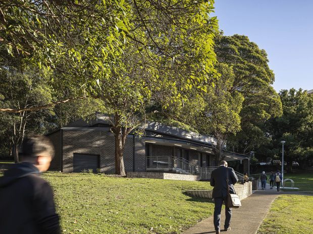 Exterior view of a low-rise dark brick building set within a leafy landscaped campus or park. A paved pedestrian walkway leads toward the building, with several people walking nearby. The building has a low, angled roof, shaded verandah-like circulation area, metal railings, and a garage-style door, surrounded by mature trees and grass.