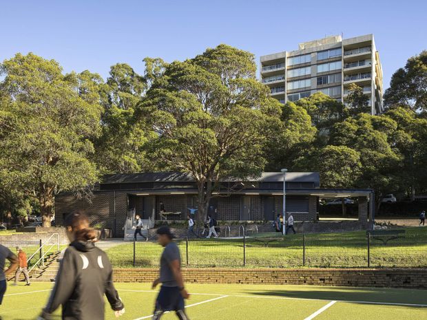 Exterior view of a low-rise contemporary brick building beside a fenced outdoor sports court, with mature trees surrounding the site and a taller multi-storey apartment building visible behind it. People are using the court and walkway in the foreground.