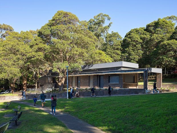 Exterior view of a low-rise modern brick building set within a leafy landscaped park or institutional grounds. The building has a flat roof with a raised box-like roof volume, dark metal roof and fascia elements, extensive brick walls, dark-framed windows, and a covered entrance or verandah supported by slender posts. A paved path, lawn, benches, mature trees, and several people surround the building.