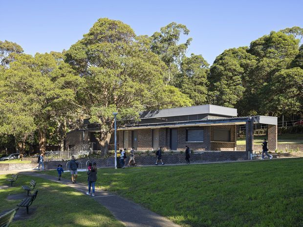 Exterior view of a low-rise contemporary brick building set within a leafy landscaped site. The building has a flat roof with projecting roof planes, dark brick walls, horizontal windows, and a covered entrance or walkway. People and benches are visible across the lawn, with mature trees forming a dense backdrop.