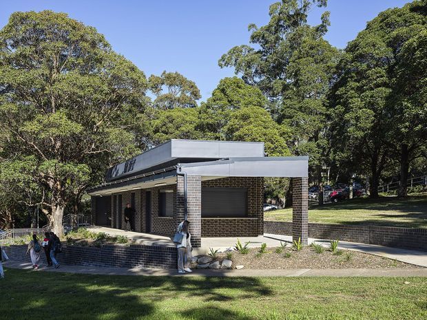 A low, single-storey contemporary brick building set within a leafy landscaped site. The building has dark brown brick walls, broad flat roof forms with pale metal fascia, deep eaves, and large shaded window openings. Paved paths and low brick retaining walls define the foreground, with mature trees surrounding the structure.